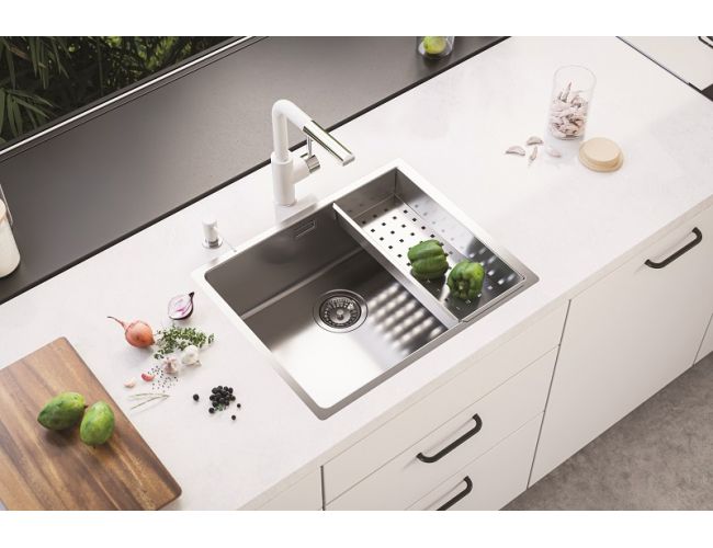 Sodobno inox pomivalno korito s perforirano odcejalno posodo, vgrajeno v bel kuhinjski pult z belo armaturo; ob njem so narezana čebula, začimbe in lesena deska, v koritu pa zelena paprika.